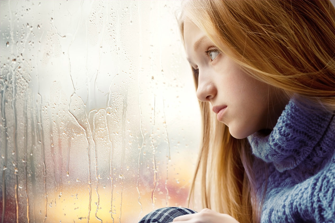 Mädchen schaut traurig aus dem Fenster; Regen läuft die Scheibe hinunter